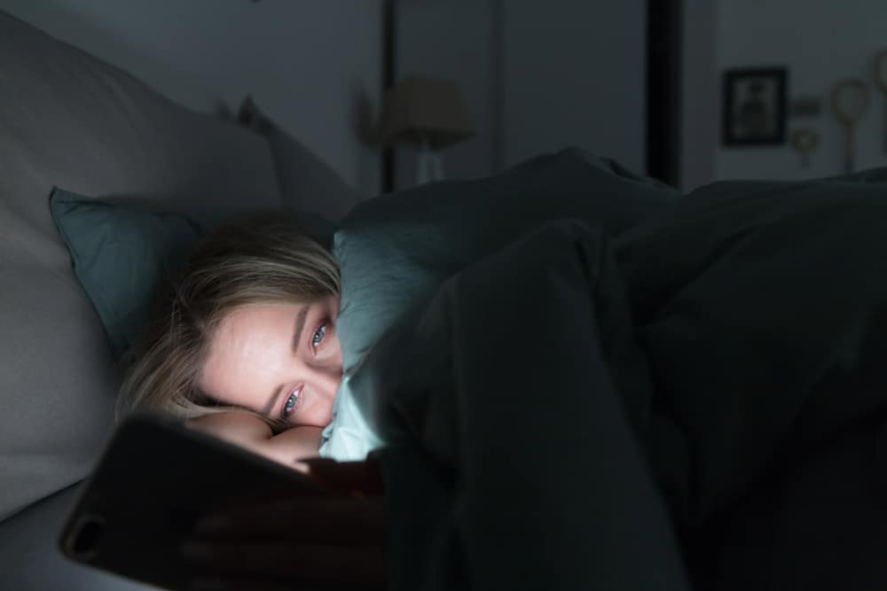 Sleeping Disorders. Tired woman lying in bed under the blanket using smartphone at late night as can not sleep.